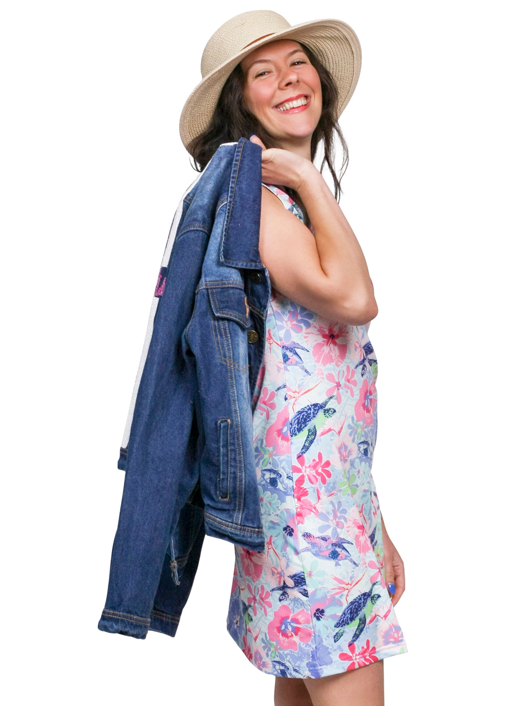 Woman smiling in a straw sun hat and sleeveless dress with a pink hibiscus and sea turtle print, carrying a denim jacket over her shoulder