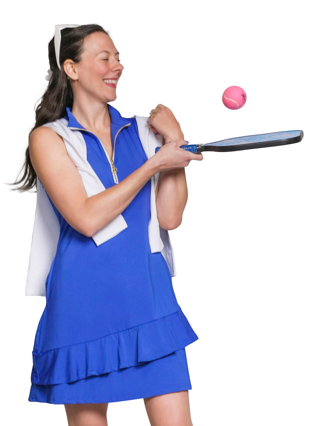 Woman in a cobalt blue sleeveless quarter-zip tennis dress with a ruffled hem, white sweater over her shoulders, hitting a ball with a paddle