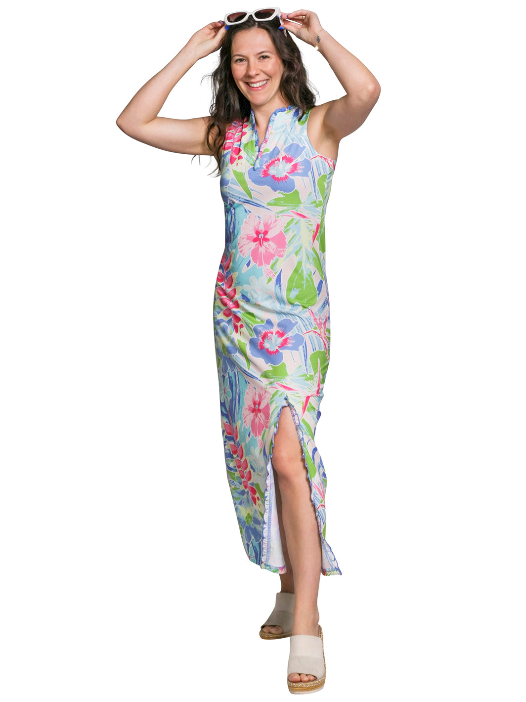 Woman smiling in a sleeveless tropical floral maxi dress with a ruffle-trimmed front slit, white sunglasses on her head and cream sandals